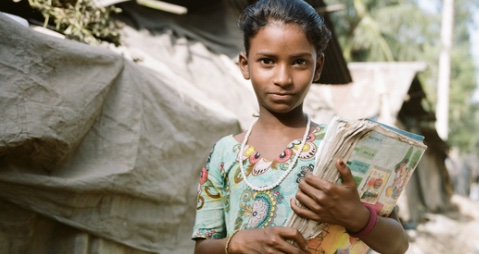 A child with short hair wearing a bead necklace and brightly colored shirt holds a stack of school papers.