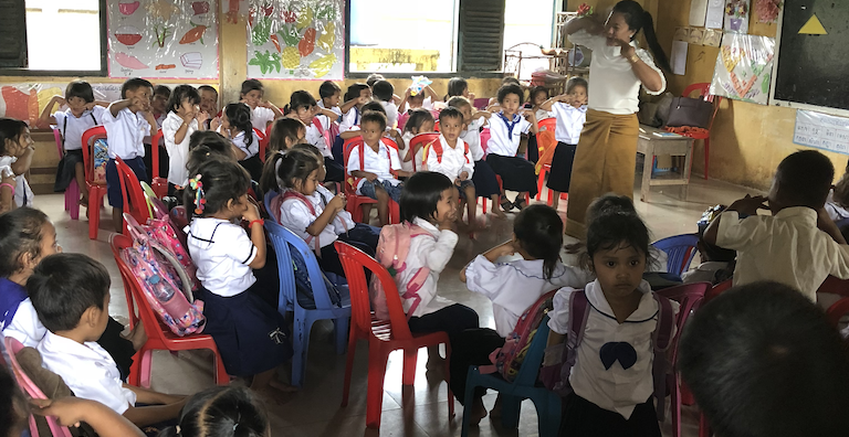 A lone teacher stands before a large class of young children in school uniforms.