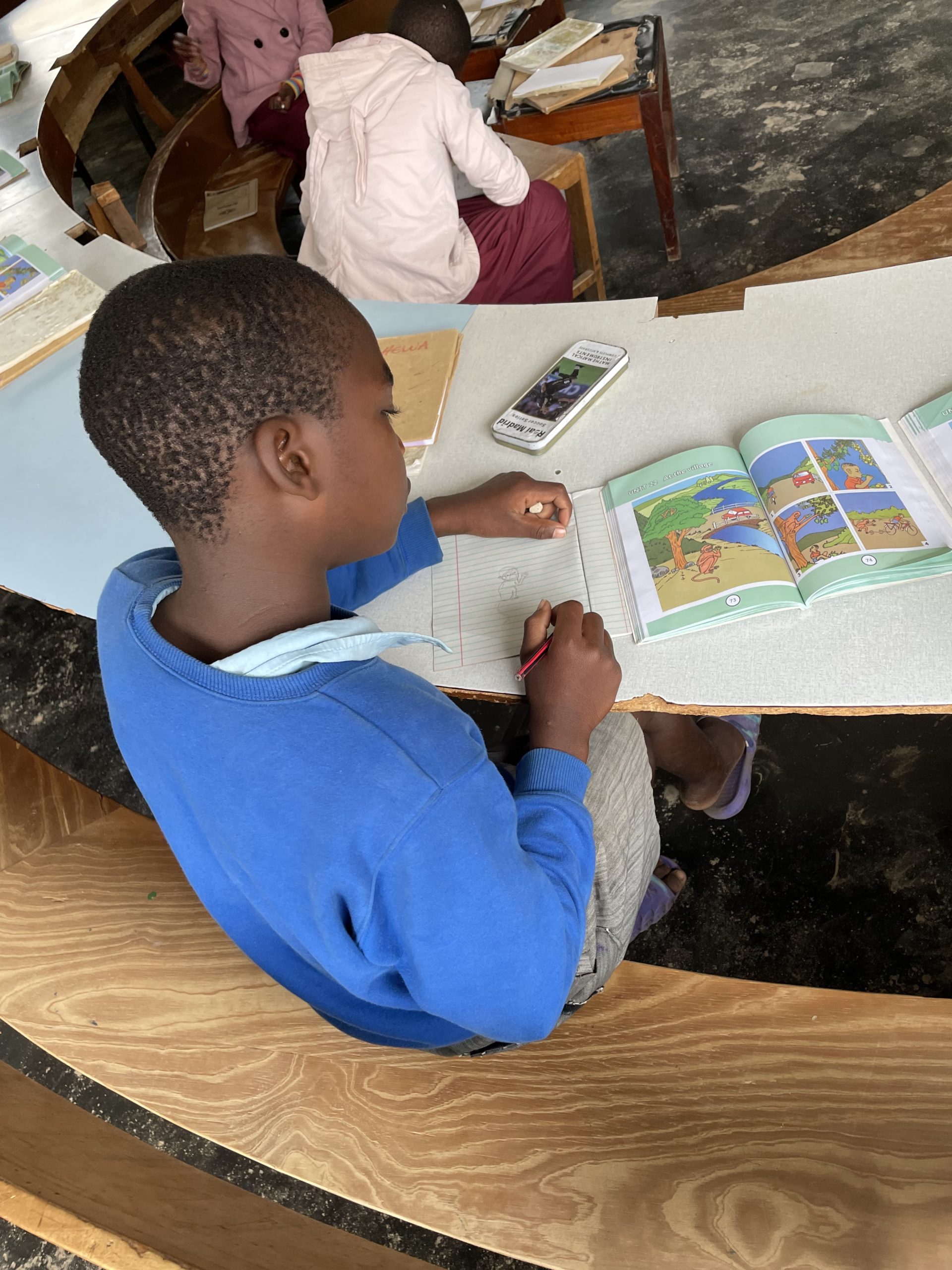 A child sits in a classroom with a book opened to show brightly colored pages. Other learners are seated nearby.