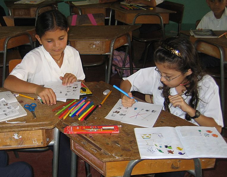 Two children seated at desks working and drawing on workbooks. A few more schoolchildren at other desks are partly visible behind them.