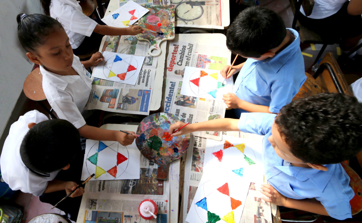 Children in white and blue school uniforms are seated at desks painting triangles