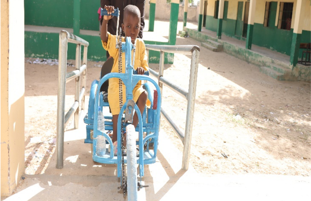 A boy on a tricycle operated with hand pedals comes up a gently sloping ramp with assistance from the man behind him.