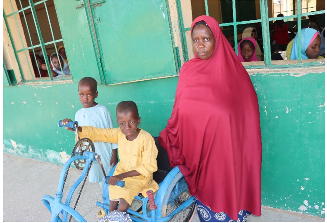 A woman in a long red hijab is standing next to two boys, one of whom rides a wheelchair configured like a tricycle but operated with hand pedals.