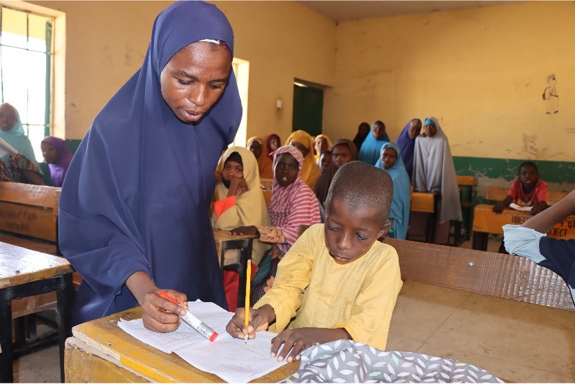A teacher in a long blue hijab stands next to a boy who is seated at a classroom desk writing on paper.