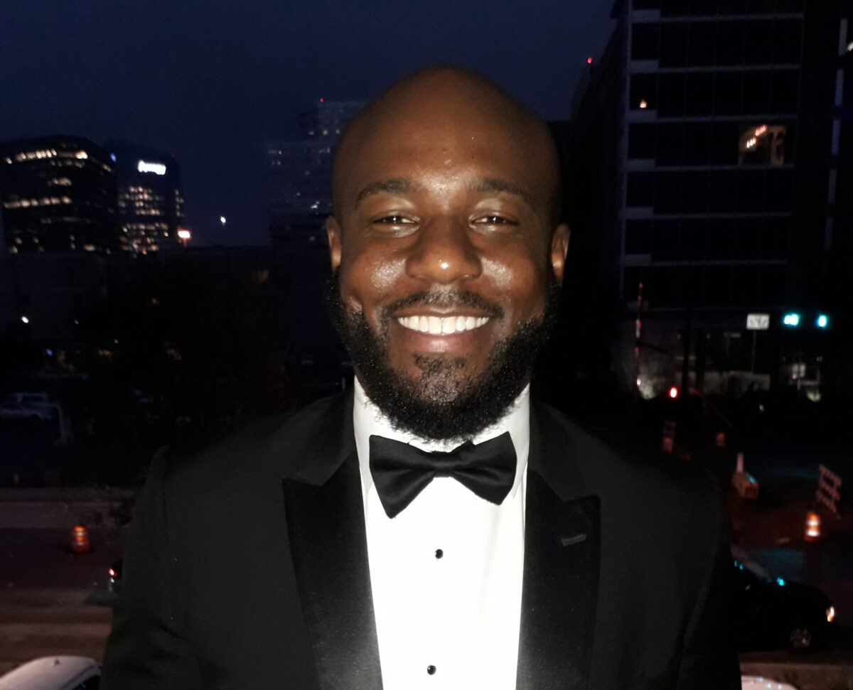 A man in a tuxedo, standing outside at night, smiles at the camera.