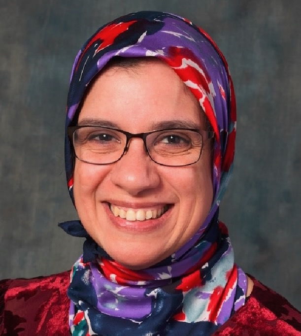 A woman wearing glasses and a bright and colorful hijab smiles at the camera.