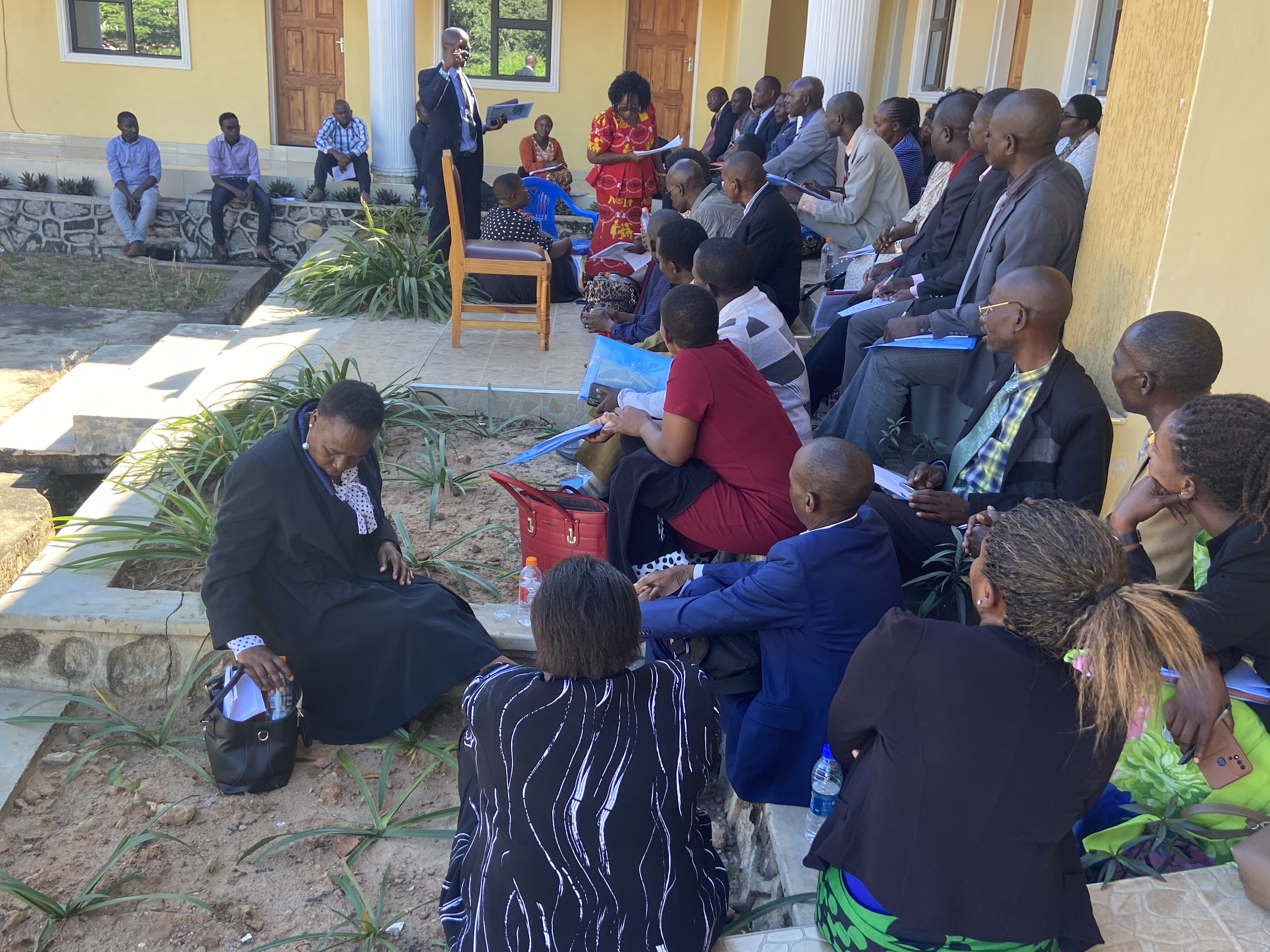 A large group of people are outside in front of a building. Most are seated on the ground, on stone walls, or on steps while others are standing. Some are interacting with each other.