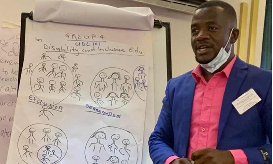 Peter Harris, with a surgical mask hanging on his chin and wearing a suit, stands next to a large sheet of paper on an easel. The paper has hand-made drawings made with a black marker. At a teacher training on inclusive education in Liberia.